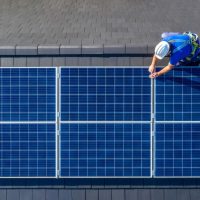 Male engineer in blue suit and protective helmet installing photovoltaic panel system. Professional electrician mounting solar module on roof. Alternative energy ecological concept